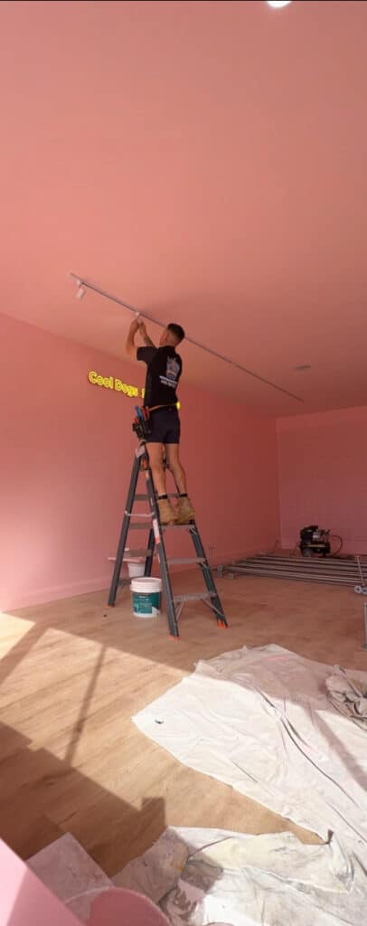 An electrician installing lights in a commercial space with pink walls.
